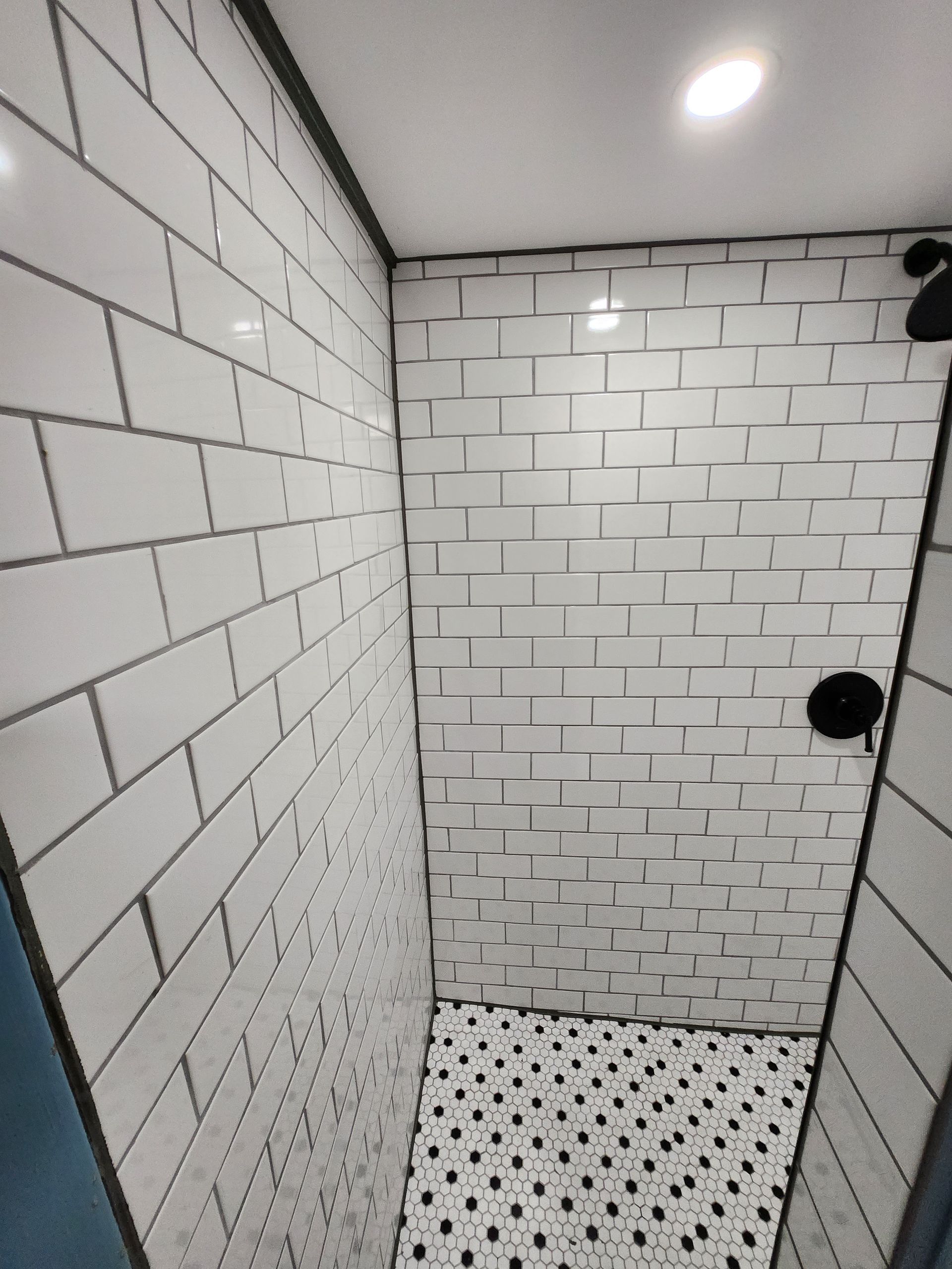 White-tiled shower with black grout. Black shower fixtures and a patterned black and white floor.
