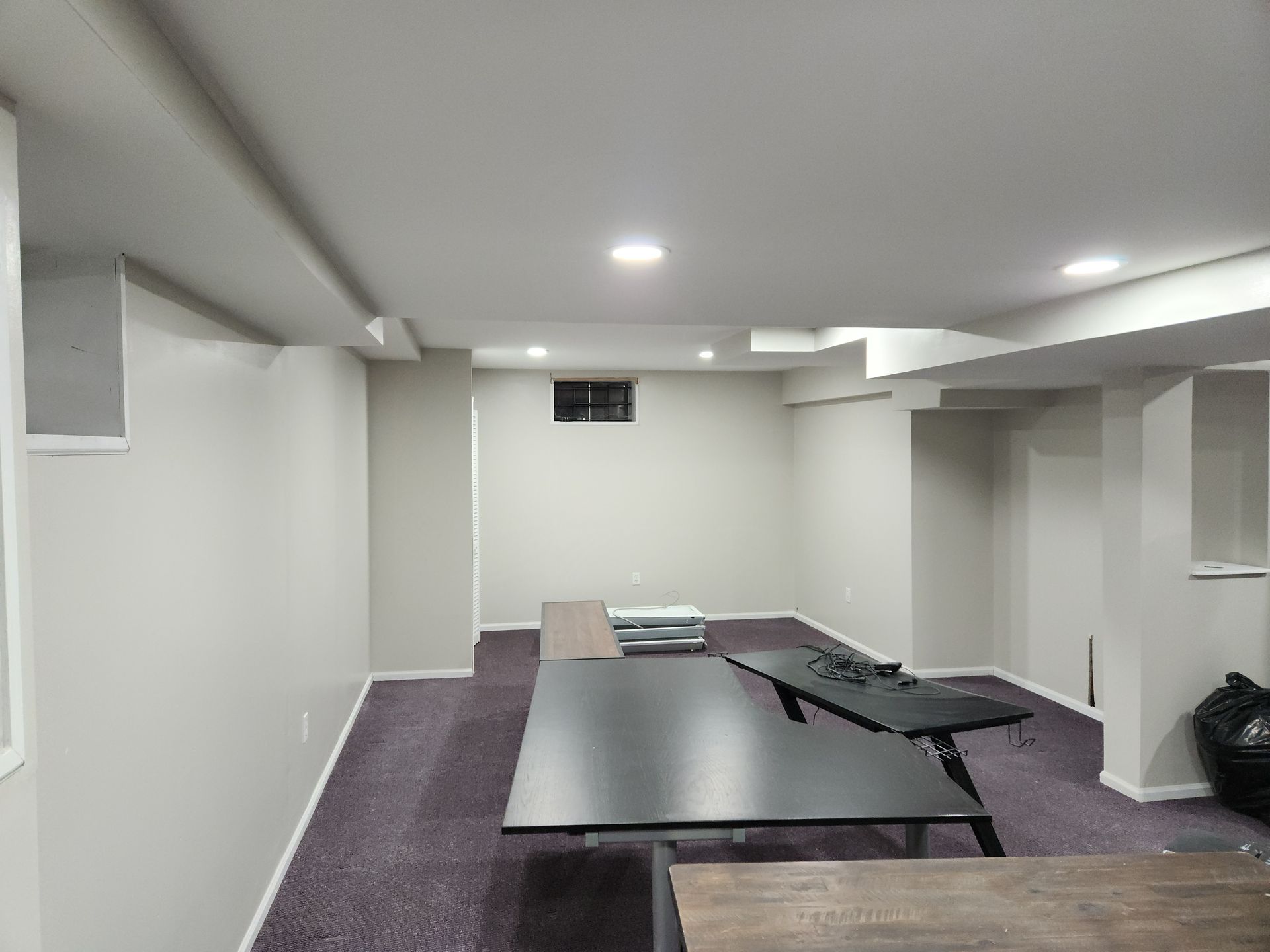 Empty, brightly lit basement room.  Dark tables on purple carpet, white walls, support beams, and ceiling lights.