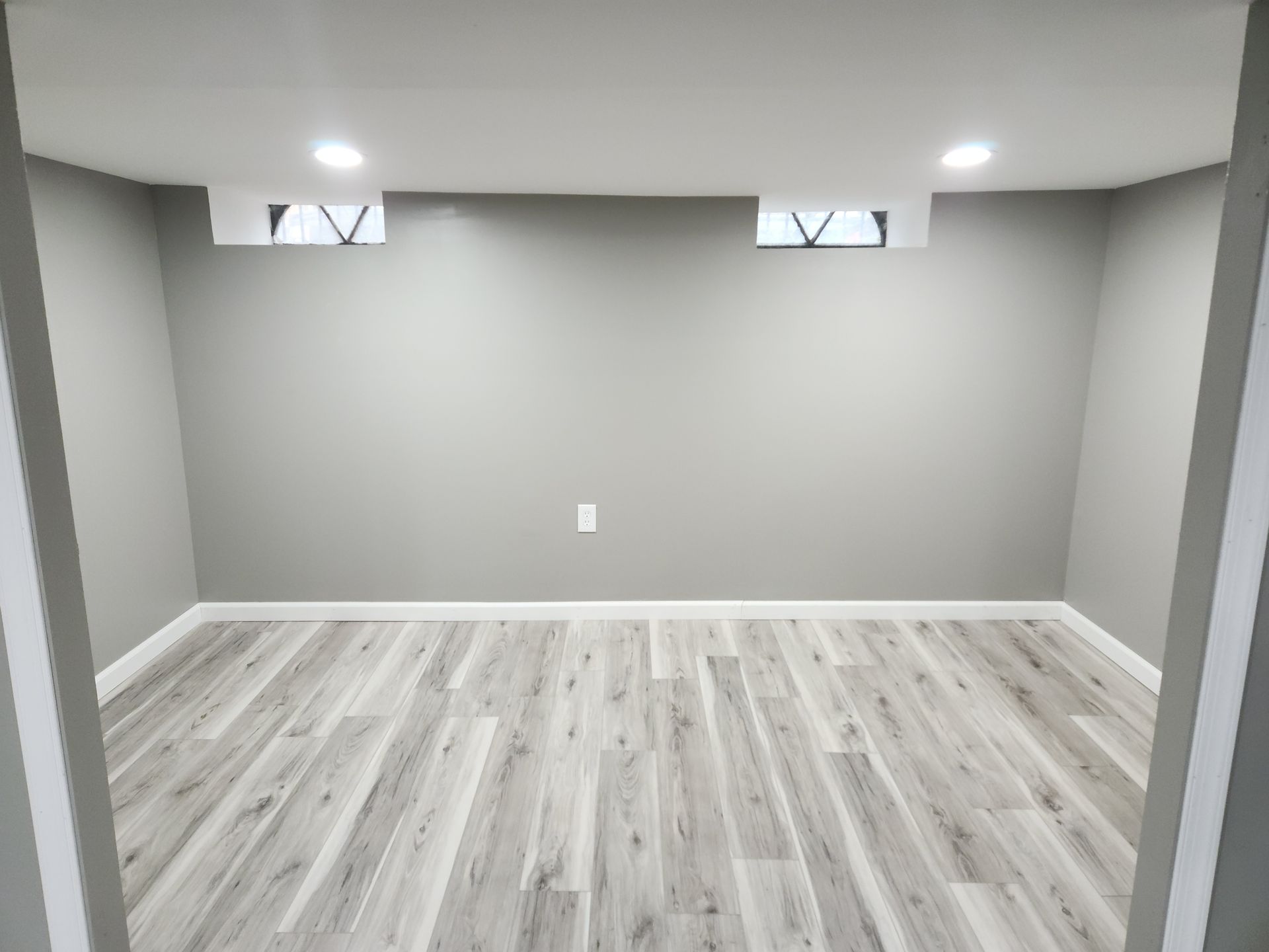 Empty finished basement room with gray walls, light wood floors, and small high windows.