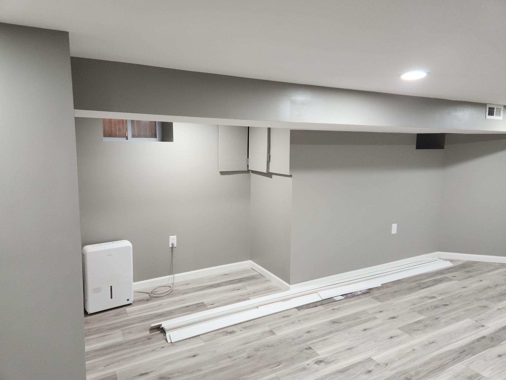 Empty gray-painted basement with wood-look flooring, exposed beams, and a dehumidifier against a wall.