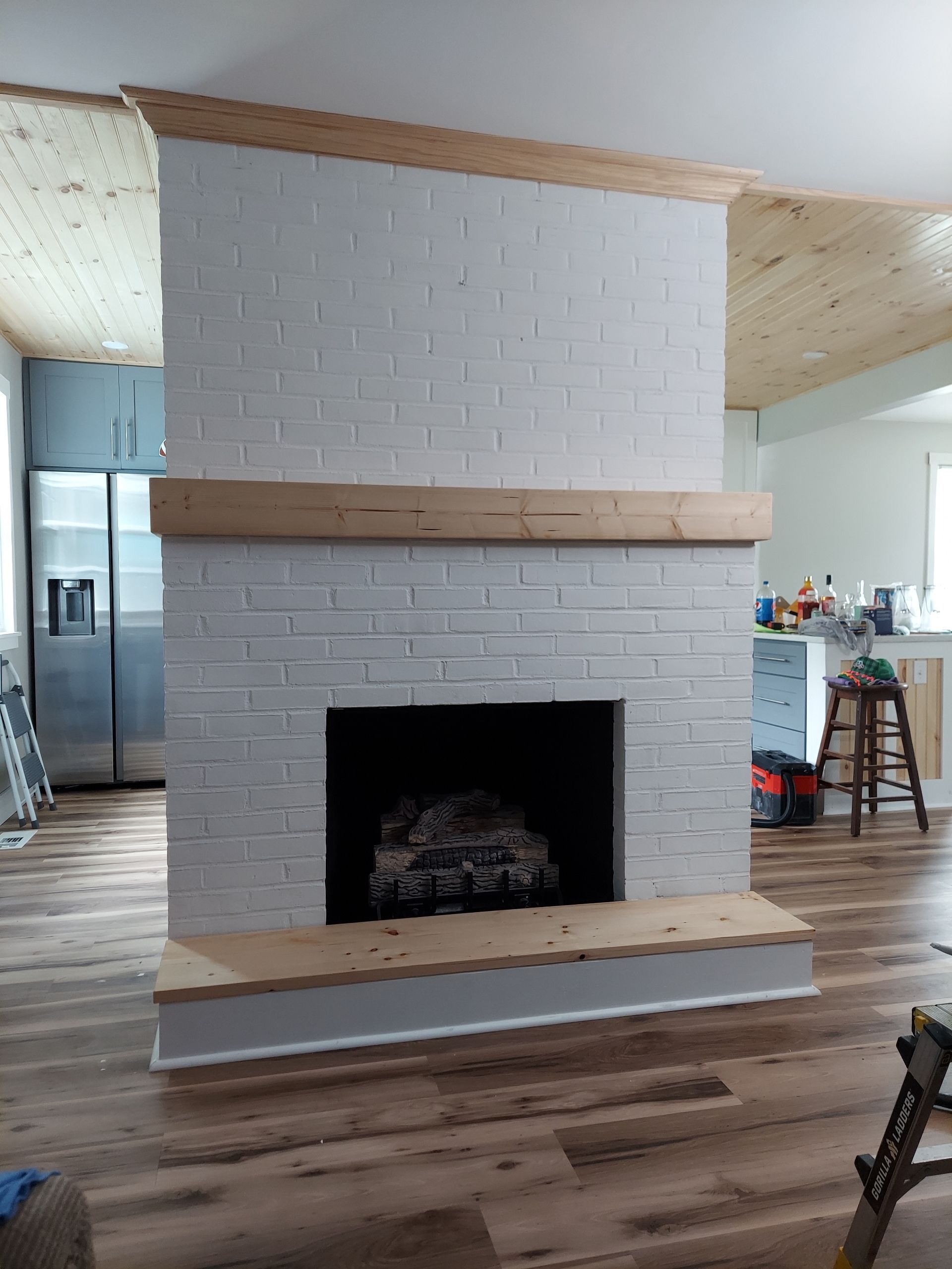 White-painted brick fireplace with a wooden mantel and hearth, in a room with wood flooring and a plank ceiling.