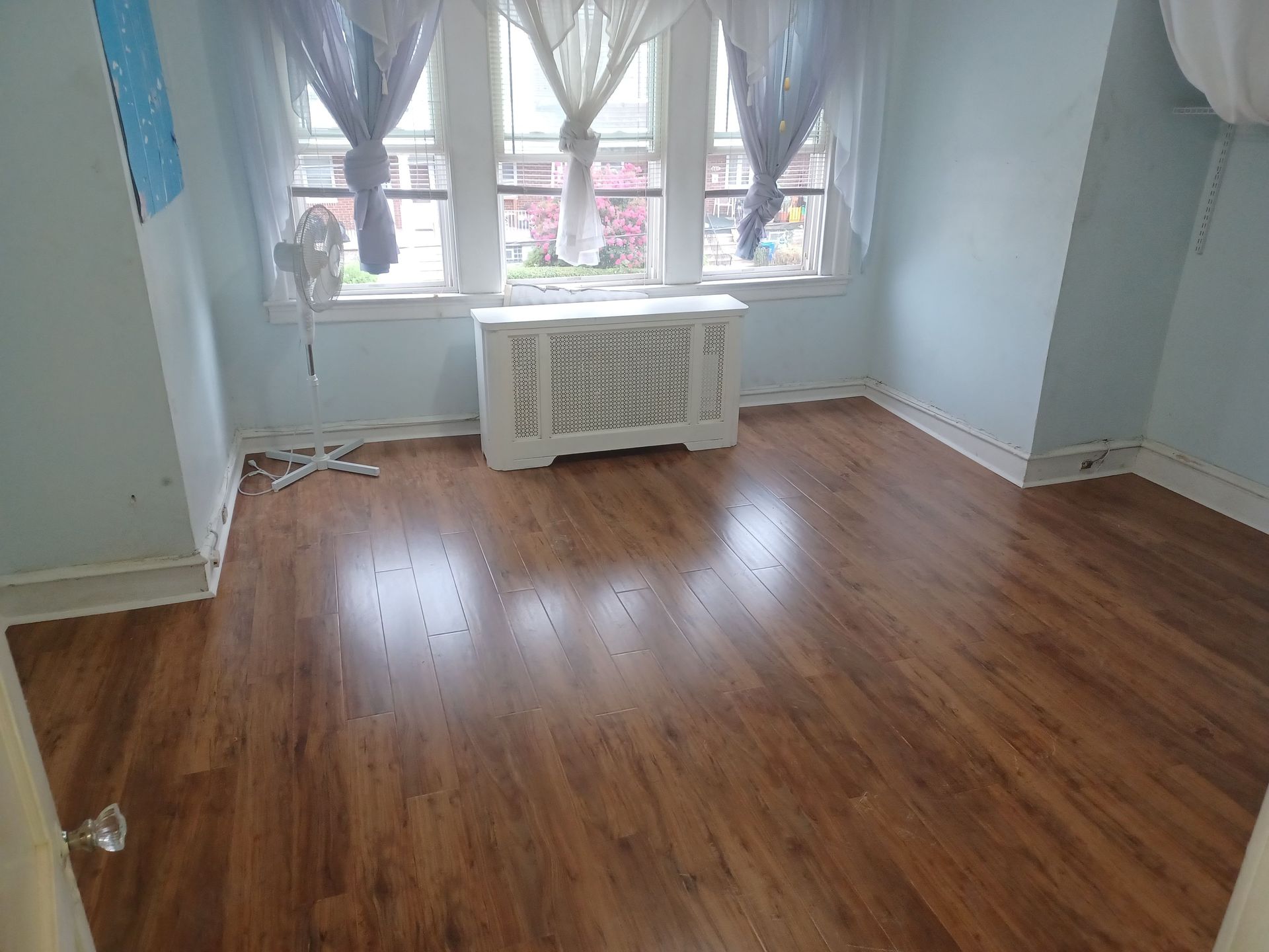 Empty room with wooden floor, window, and white radiator. Blue walls and sheer curtains.