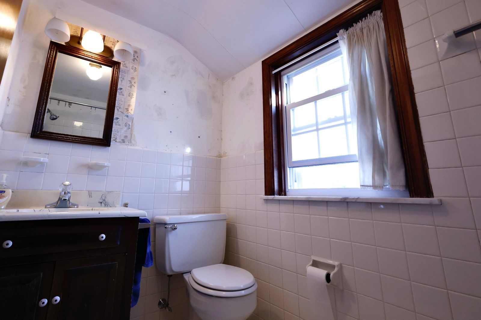 Bathroom with white tile walls, a toilet, a window with curtains, and a dark-framed mirror.