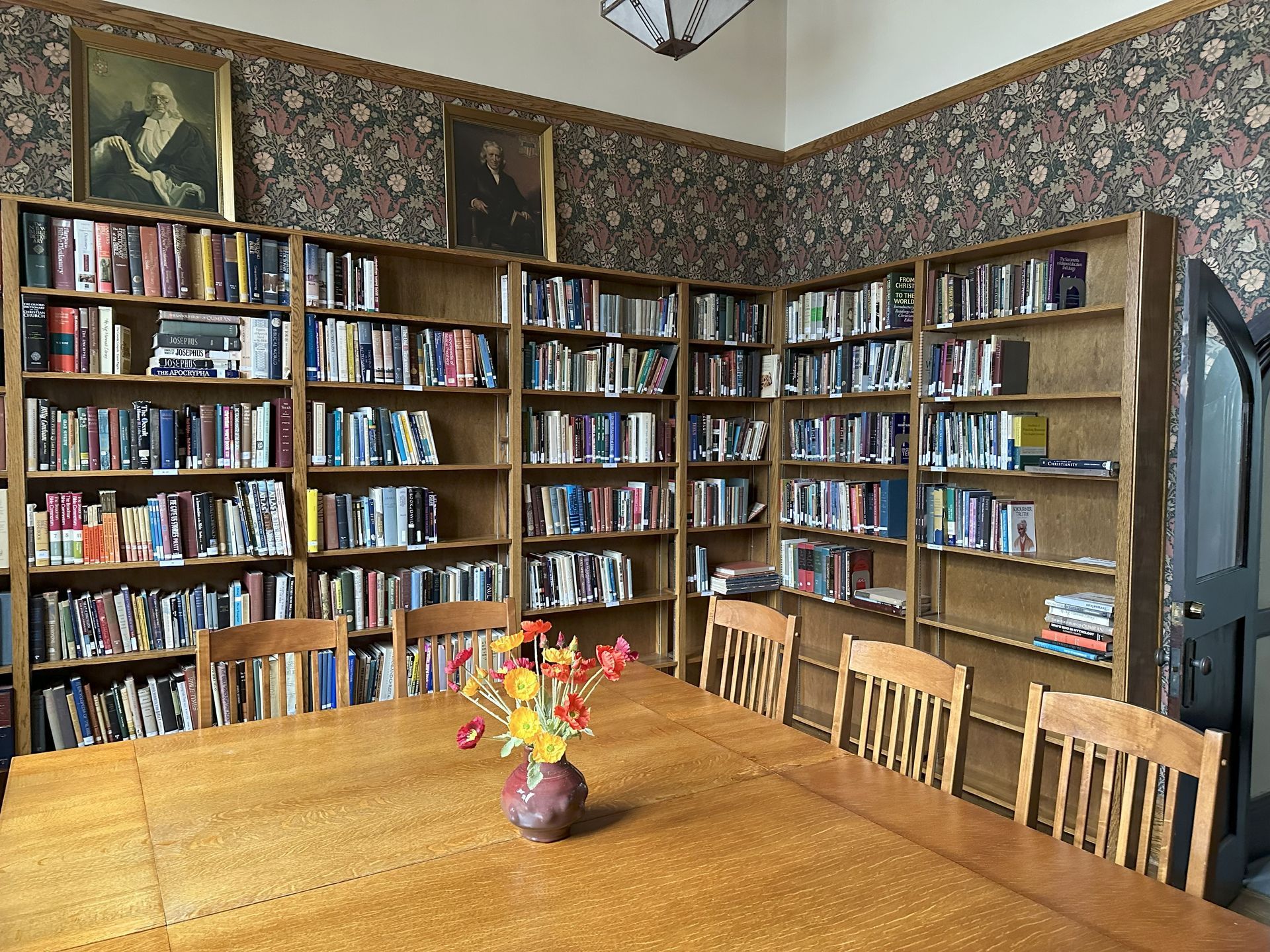Library corner with wooden shelves full of books, large table, chairs, floral wallpaper, artwork, and vase.