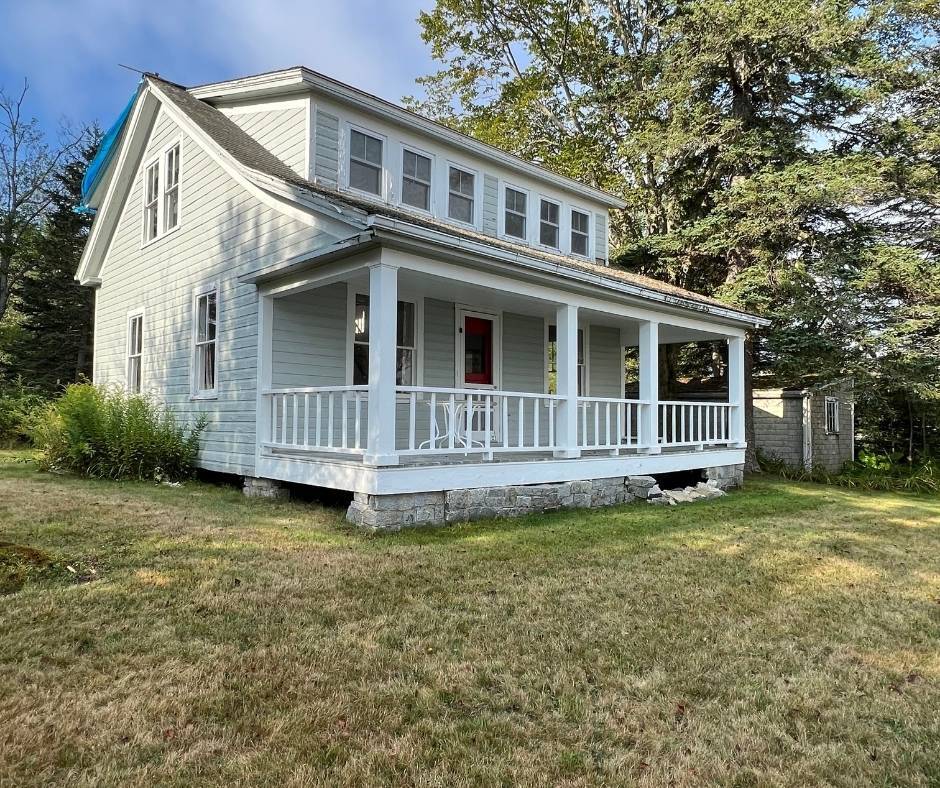 A white house with a large porch is sitting on top of a lush green field.