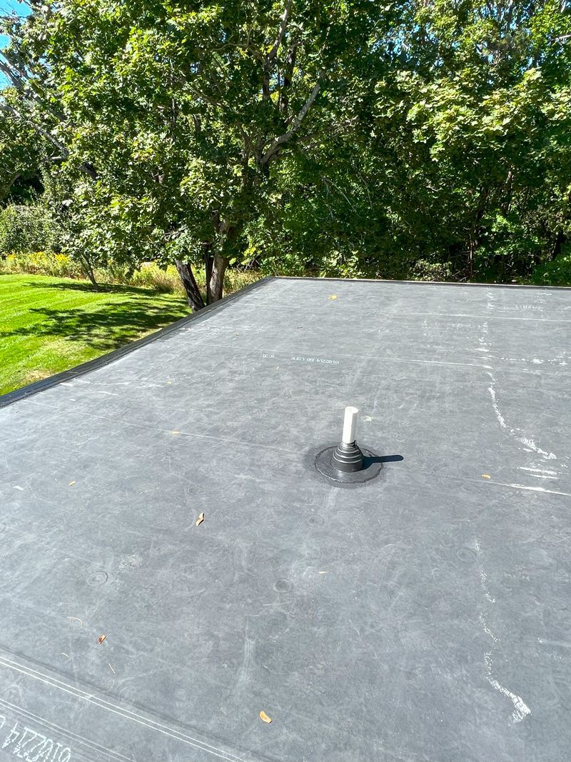 A black roof with a white pipe on it and trees in the background.