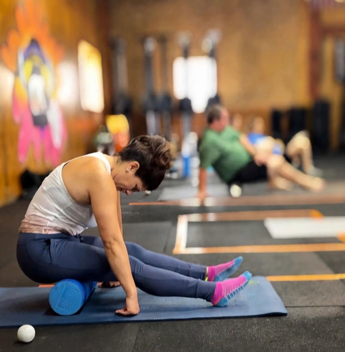 Person stretching on a blue mat in a yoga studio, using a foam roller and ball for recovery exercises