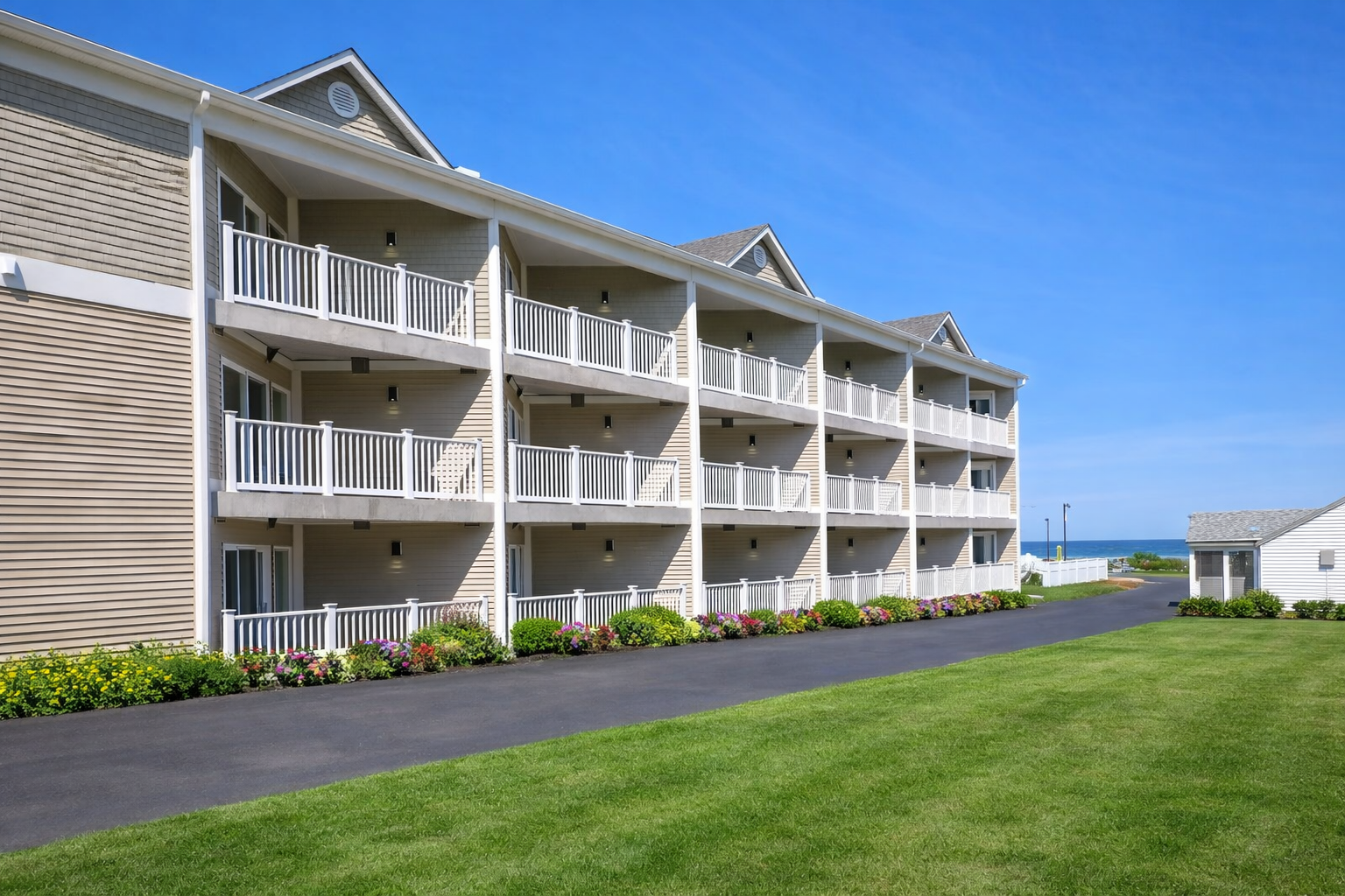 Three-story tan hotel building with balconies facing a lawn and a paved driveway under a clear blue sky.