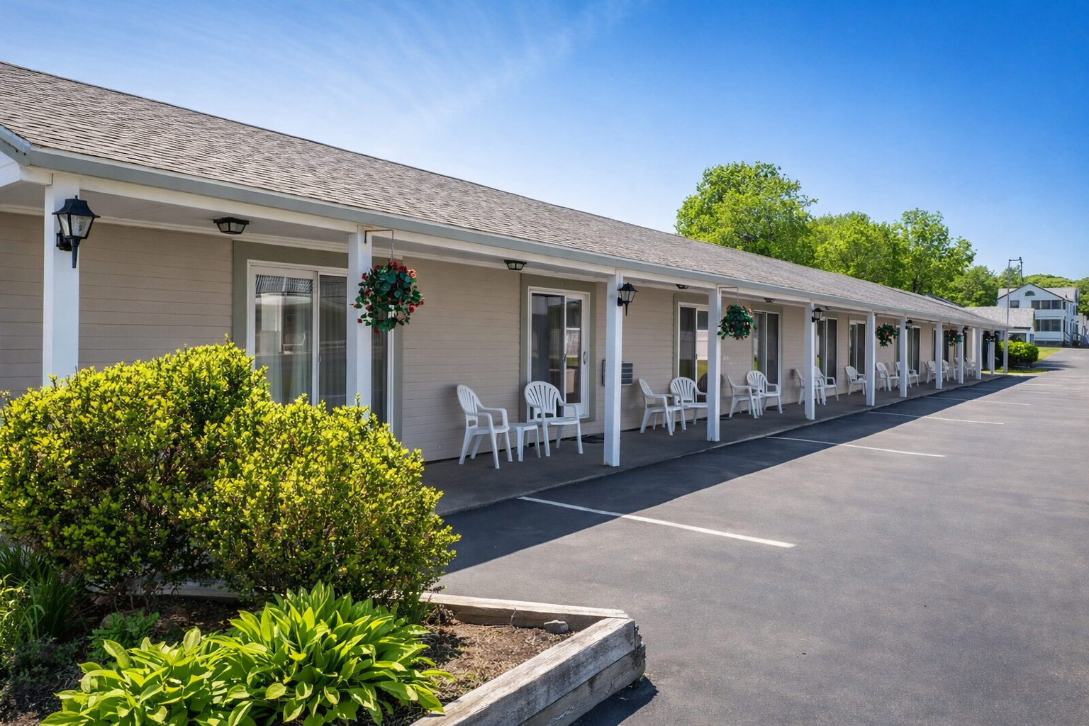 A beige, single-story hotel building with white pillars, outdoor seating, and a paved parking lot under a clear blue sky.