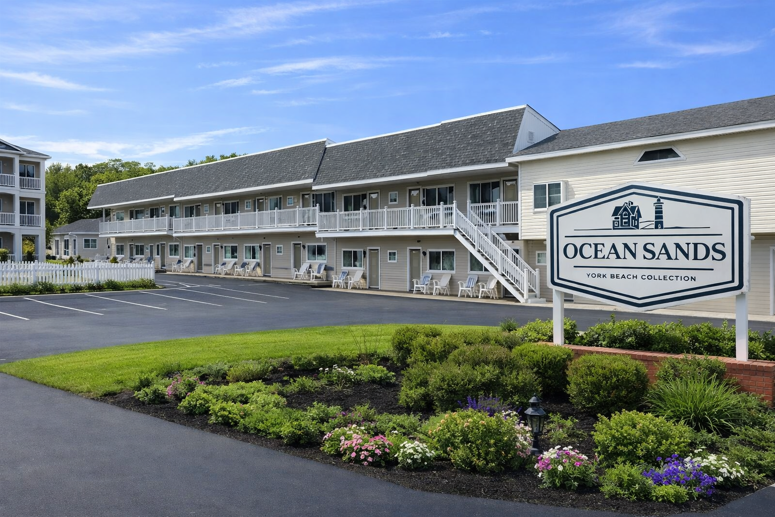 A two-story hotel named Ocean Sands at York Beach under a blue sky, with white balconies, parking lot, and landscaping.