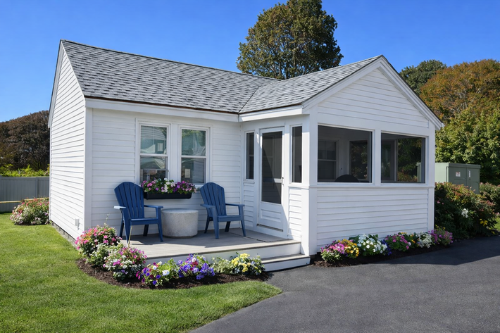 A small white cottage with a porch, blue chairs, and colorful flower beds on a sunny day.