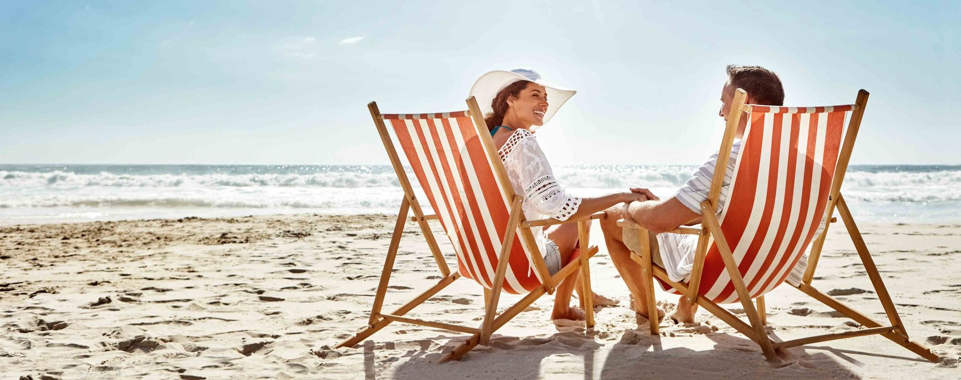 A couple relaxes in red and white striped wooden deck chairs on a sandy beach, looking toward the ocean.