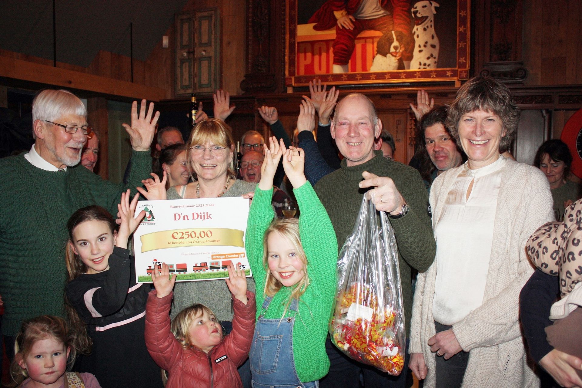 Een groep viert feest met een grote cheque en een zak snoep. Mensen steken hun armen omhoog en lachen, binnen.