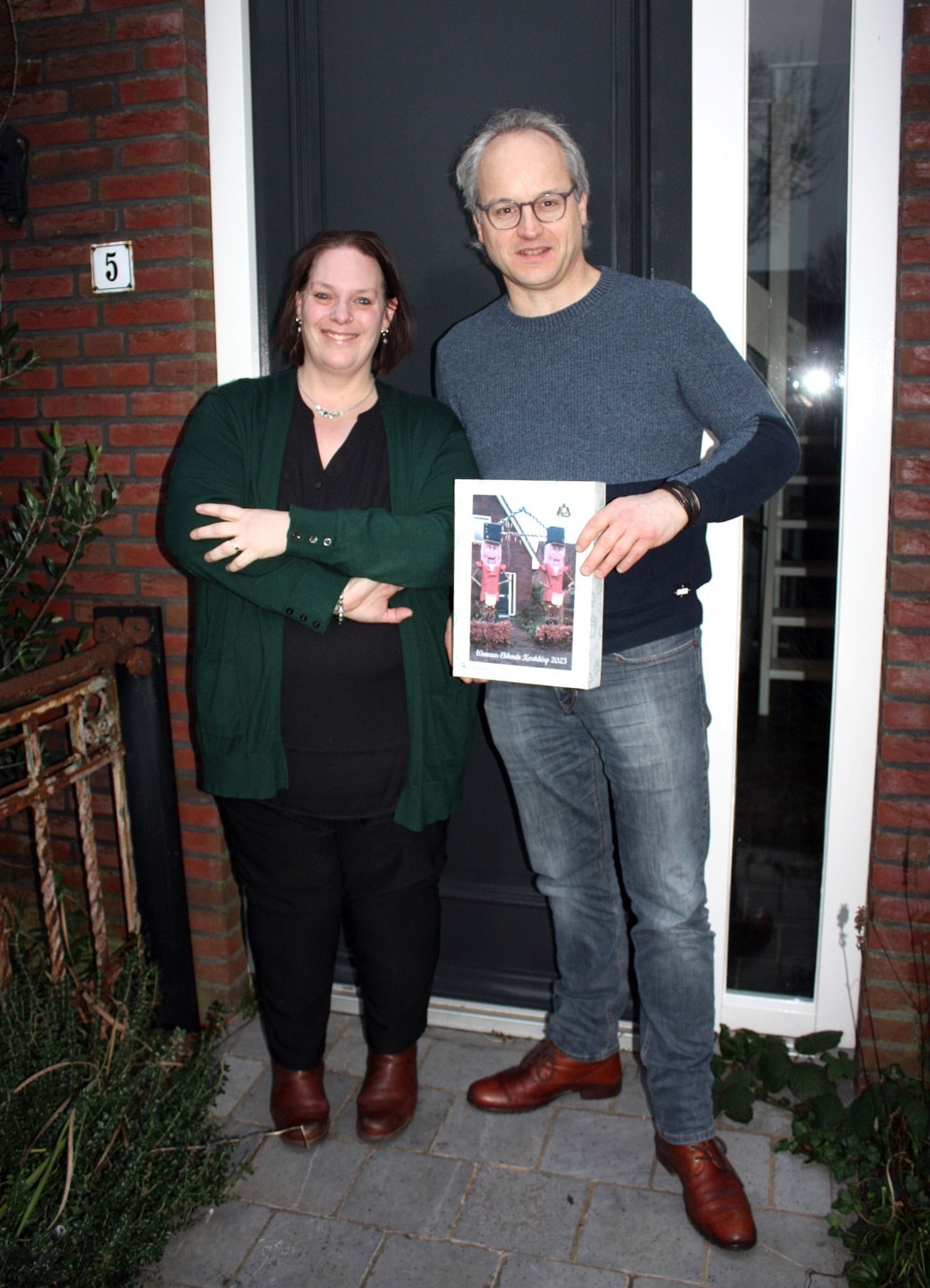 Twee mensen staan voor een deur, een van hen houdt een krant vast. De vrouw draagt zwart en een groene trui. De man draagt blauw en een spijkerbroek.