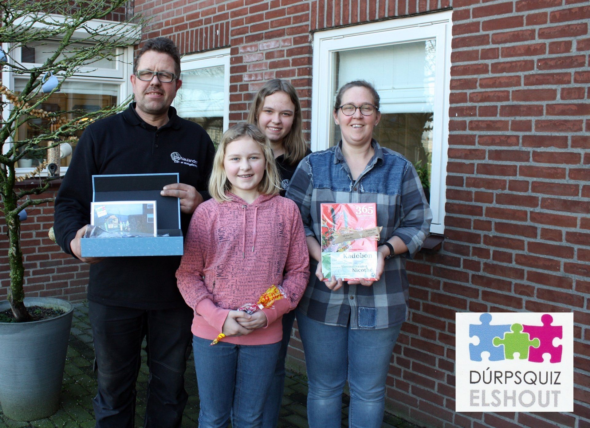 Vier mensen staan ​​buiten een bakstenen gebouw met cadeaus in hun handen. Een man heeft een blauwe doos vast, een ander een cadeau ingepakt in roze.