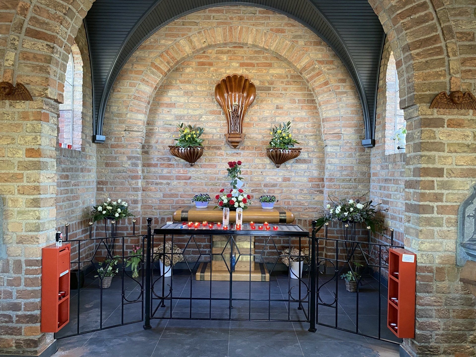 Kleine bakstenen kapel met gewelfd plafond, altaar, kaarsen en bloemen.