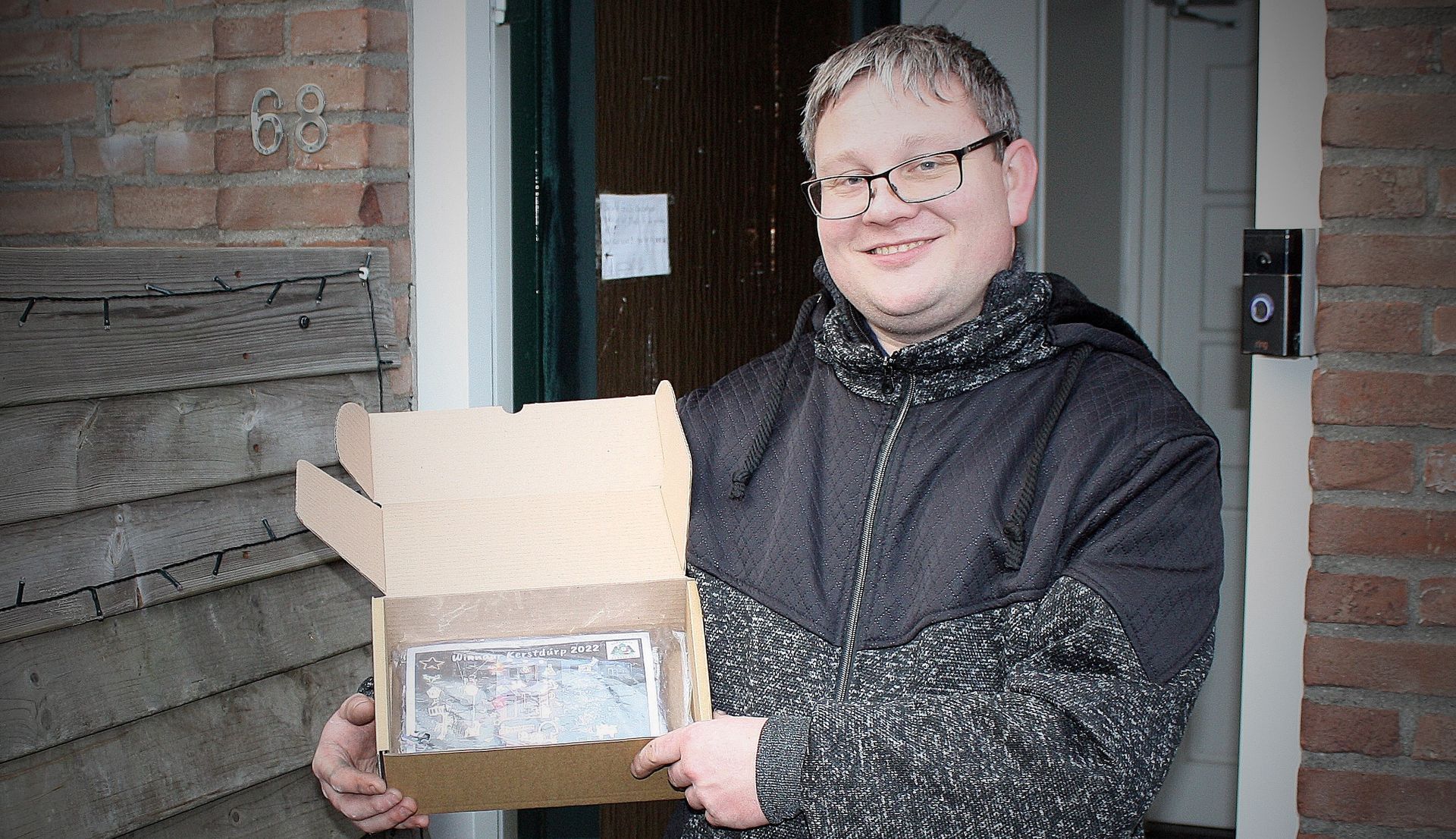 Een man houdt een kartonnen doos open en staat lachend voor een deur. Buitenopname, bakstenen gebouw, doos met inhoud.