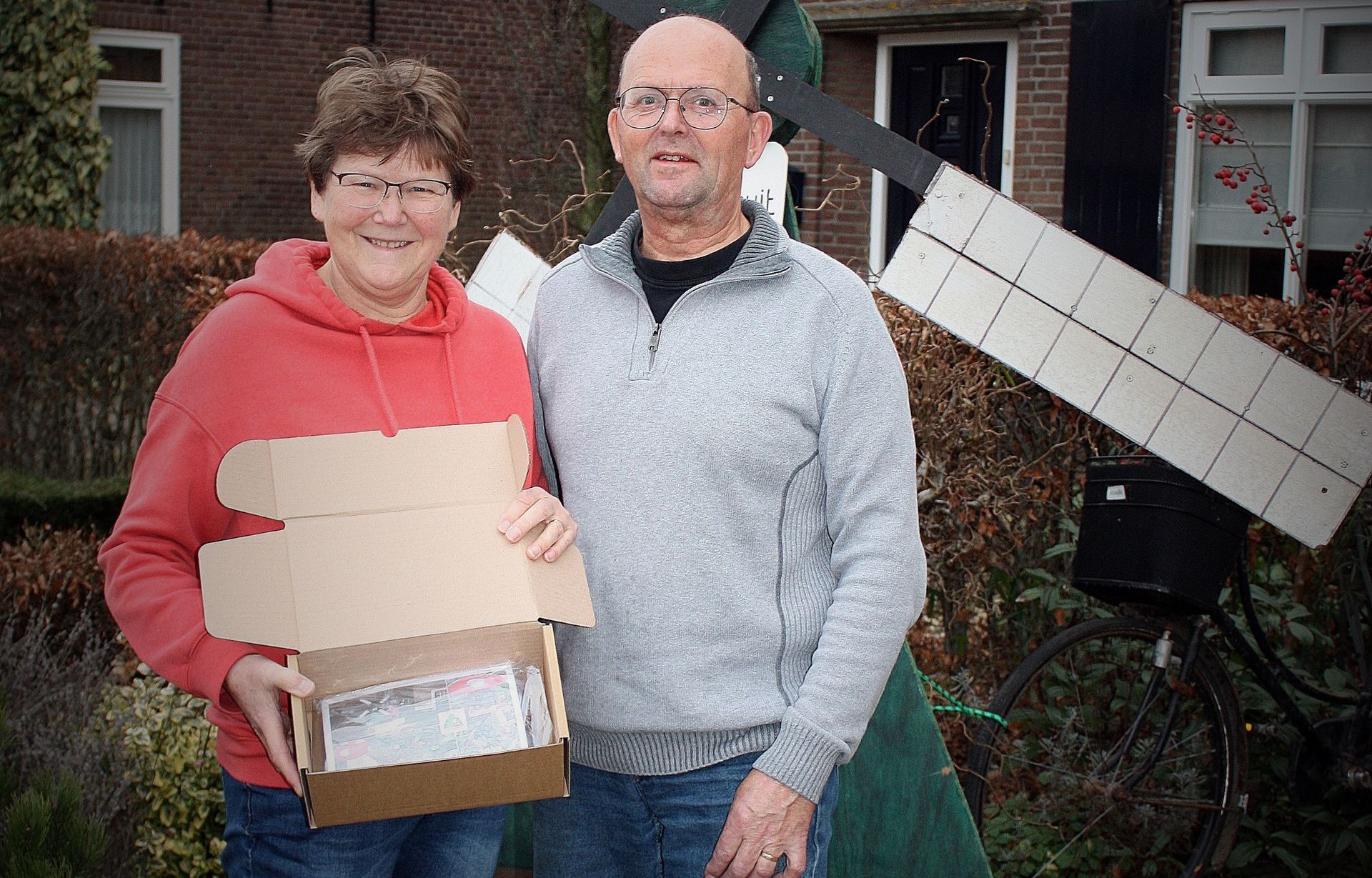 Een stel met een open kartonnen doos staat buiten bij een windmolen. De vrouw draagt ​​een rode trui, de man een grijze trui.