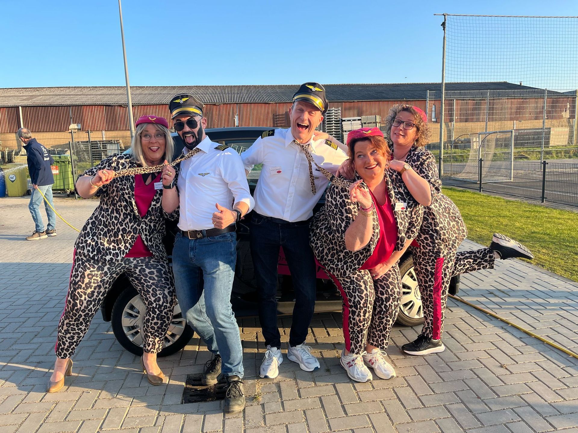 Een groep van vijf mensen poseert voor een auto. Sommigen dragen pilotenpetten en jumpsuits met dierenprint en lachen. Buiten.