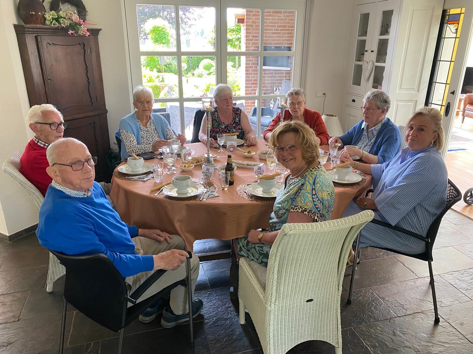 Mensen zitten aan een tafel te eten. De tafel is gedekt met servies, drankjes en eten. Een zonnige kamer.