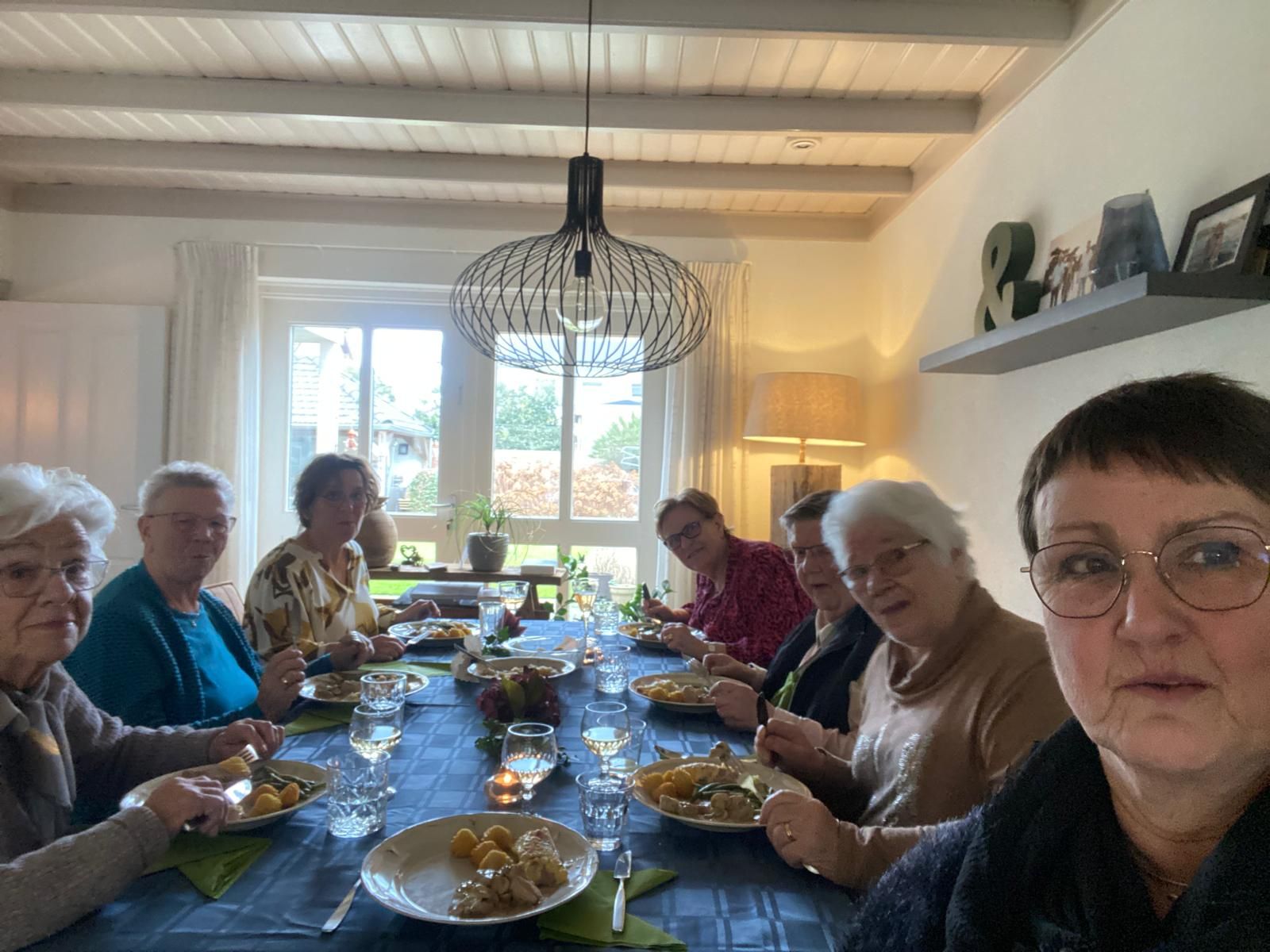 Een groep mensen zit aan een lange tafel te eten. Lichte ruimte, veel natuurlijk licht.
