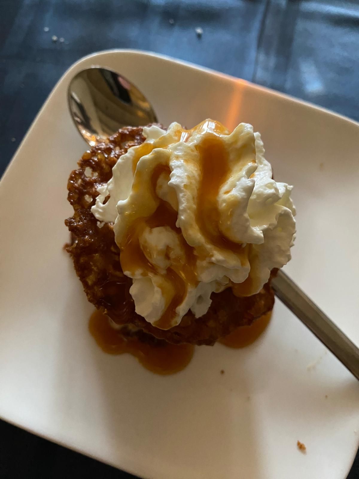 Nagerecht op een wit vierkant bord: knapperig gebak met slagroom en karamelsaus, lepel ernaast.