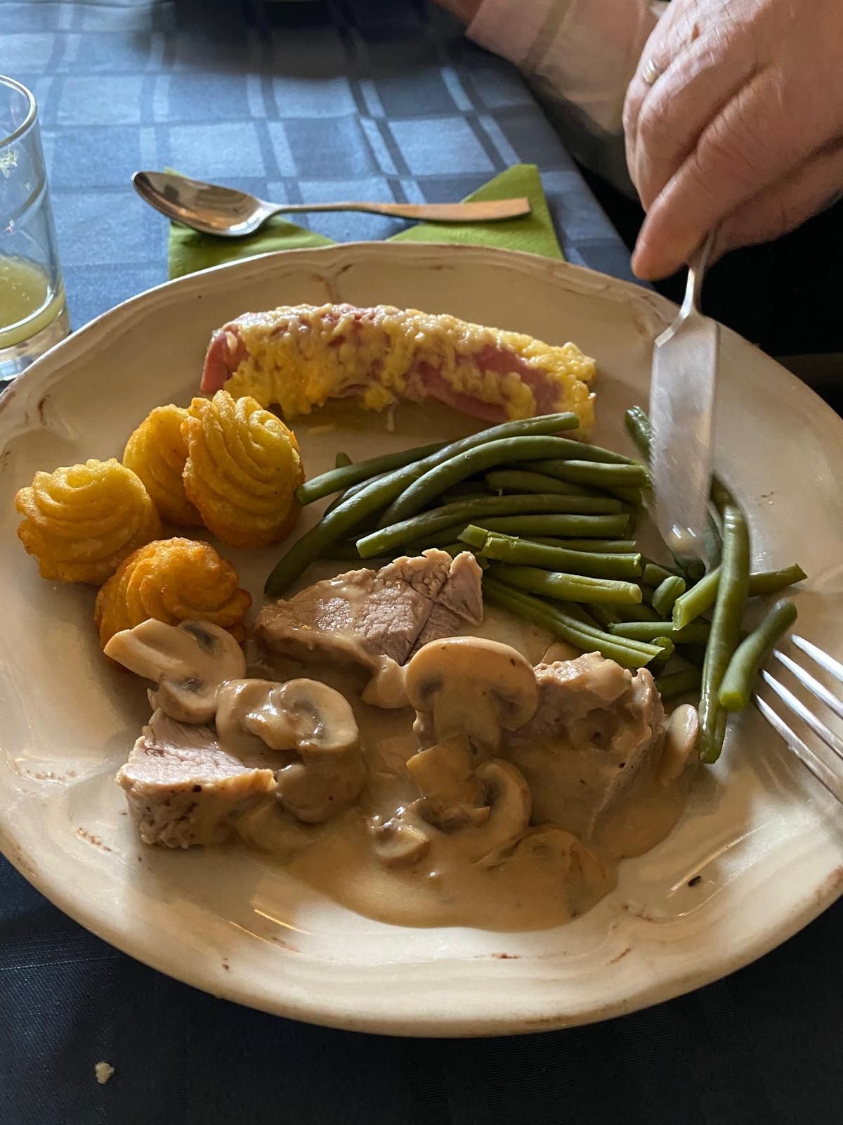 Een bord met eten: varkensvlees met champignonsaus, sperziebonen, aardappelkroketjes en gratin. Iemand gebruikt een vork en een mes.