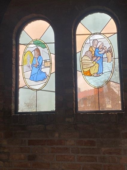 Twee glas-in-loodramen beelden religieuze taferelen uit: een figuur in blauw die knielt, figuren in interactie met elkaar, en kleurrijke achtergronden.