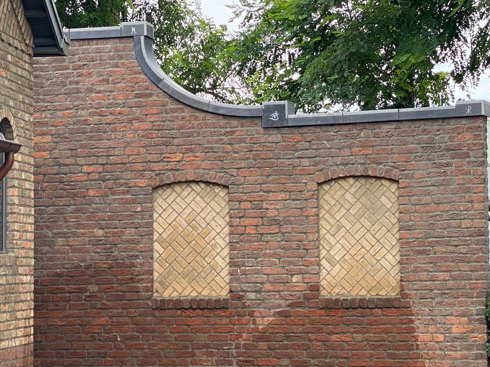 Buitenzijde van een bakstenen gebouw met twee boogvormige, dichtgemetselde ramen. Donkergrijze afdeklijst en een gebogen bovenrand.
