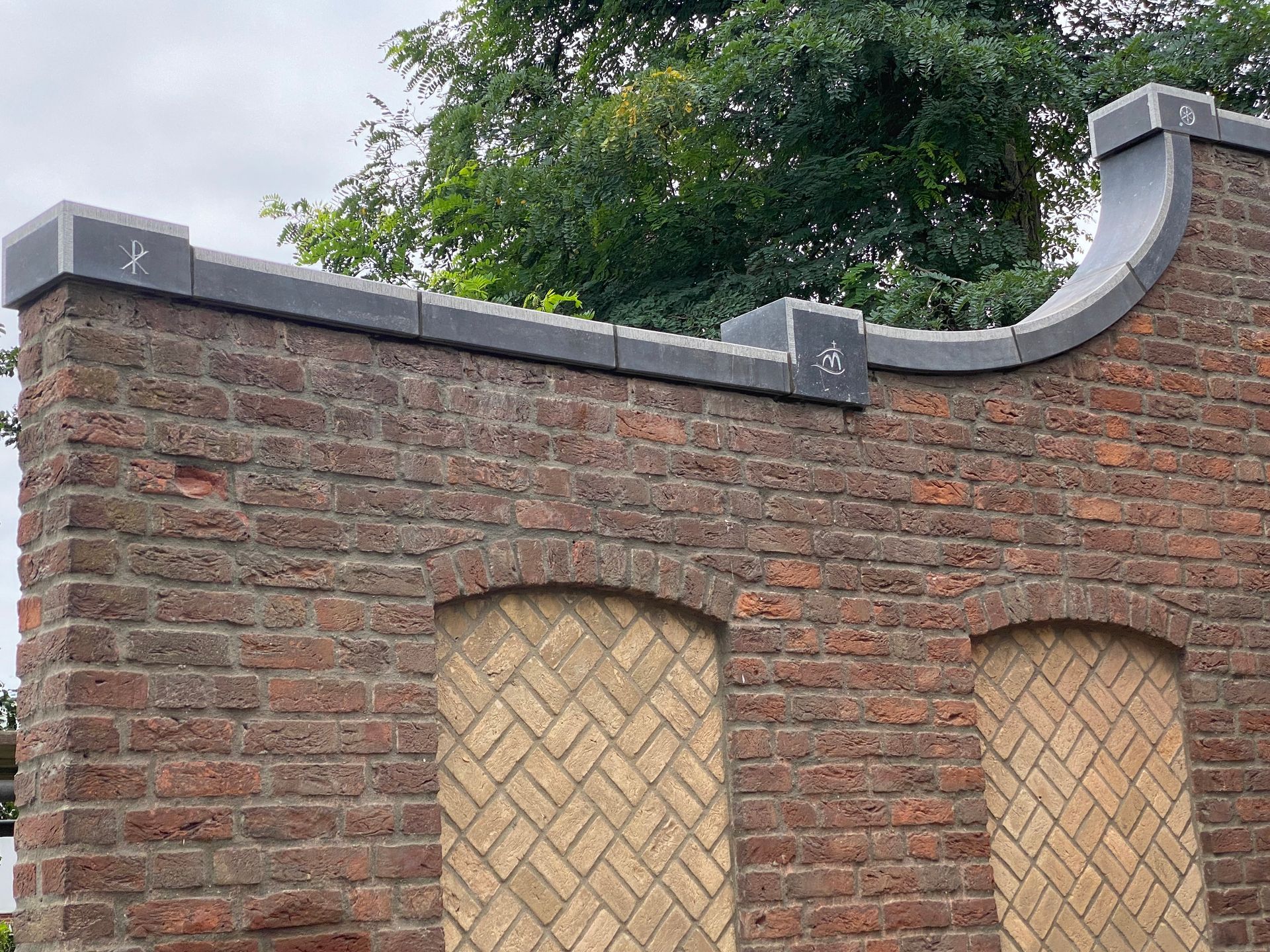 Bakstenen muur met boogvormige raamopeningen, opgevuld met bakstenen in visgraatpatroon en afgewerkt met een grijze afdekking.