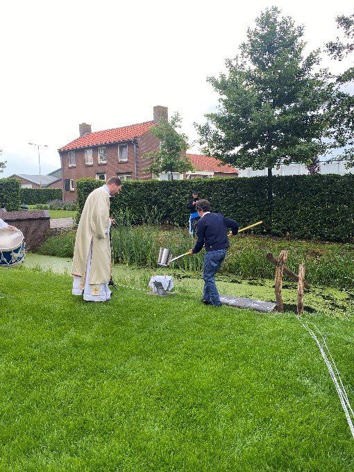 Een priester in een wit gewaad kijkt toe hoe een man een emmer leeggooit in een waterloop. Op de achtergrond zijn groen gras en gebouwen te zien.