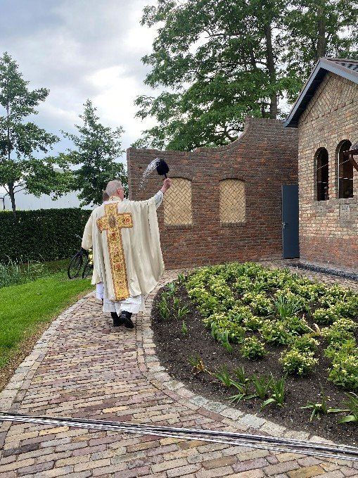 Een priester in rijkversierde gewaden neemt zijn hoed af terwijl hij over een bakstenen pad loopt, met een tuin en een bakstenen gebouw op de achtergrond.