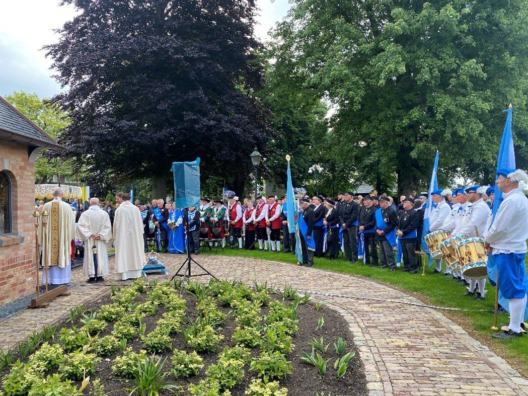 Een ceremonie met mensen in witte en blauwe uniformen nabij een gebouw, bijgewoond door anderen in een tuin.