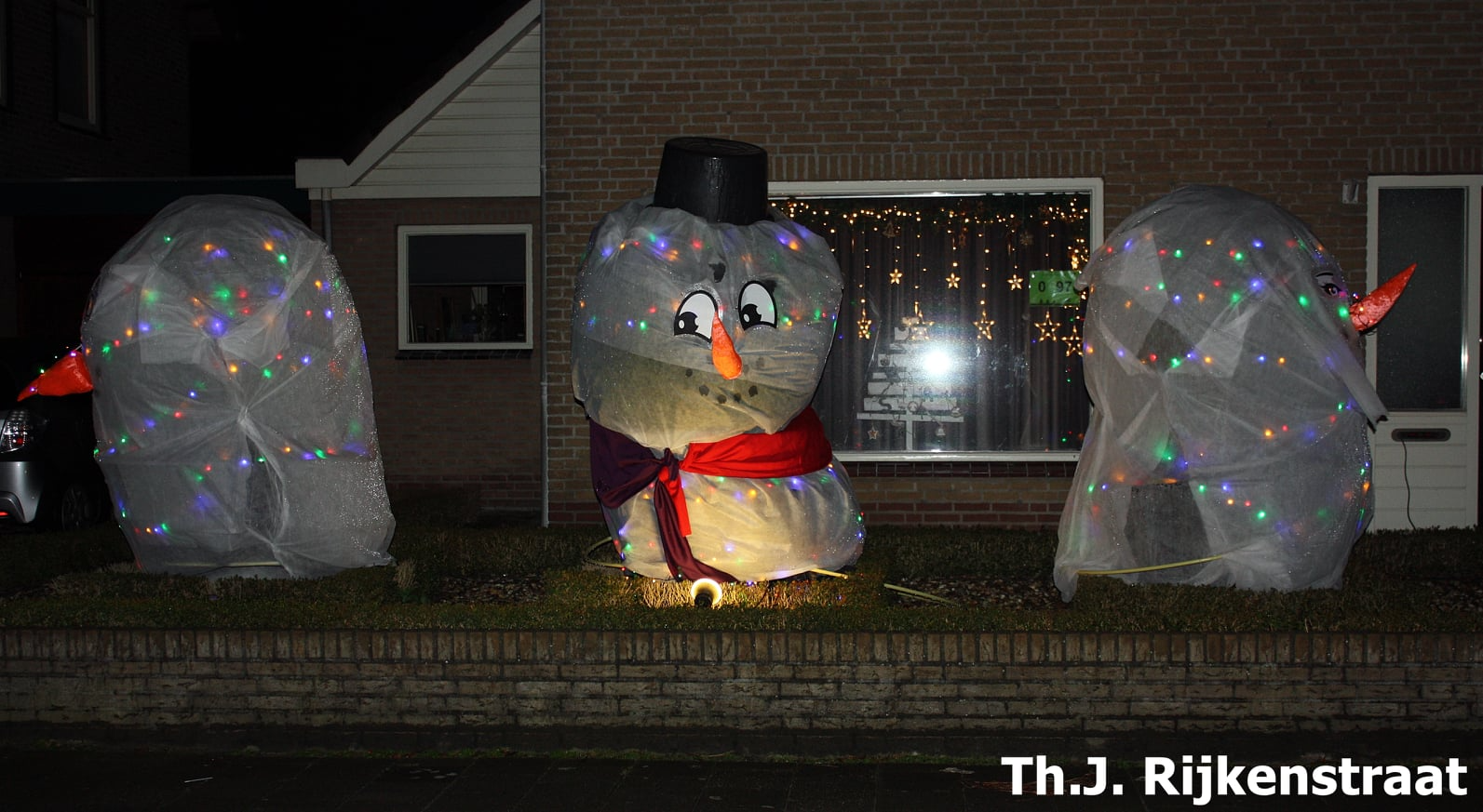 's Avonds staan er drie verlichte sneeuwpoppen in de voortuin. De middelste sneeuwpop draagt een hoge hoed en een sjaal.