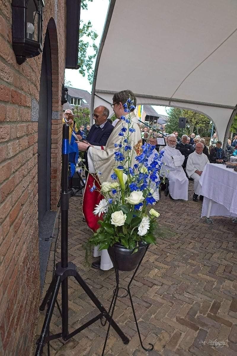 Een vrouw in een sierlijk gewaad steekt een deur aan bij een evenement in de openlucht, waar toeschouwers onder een luifel zitten.