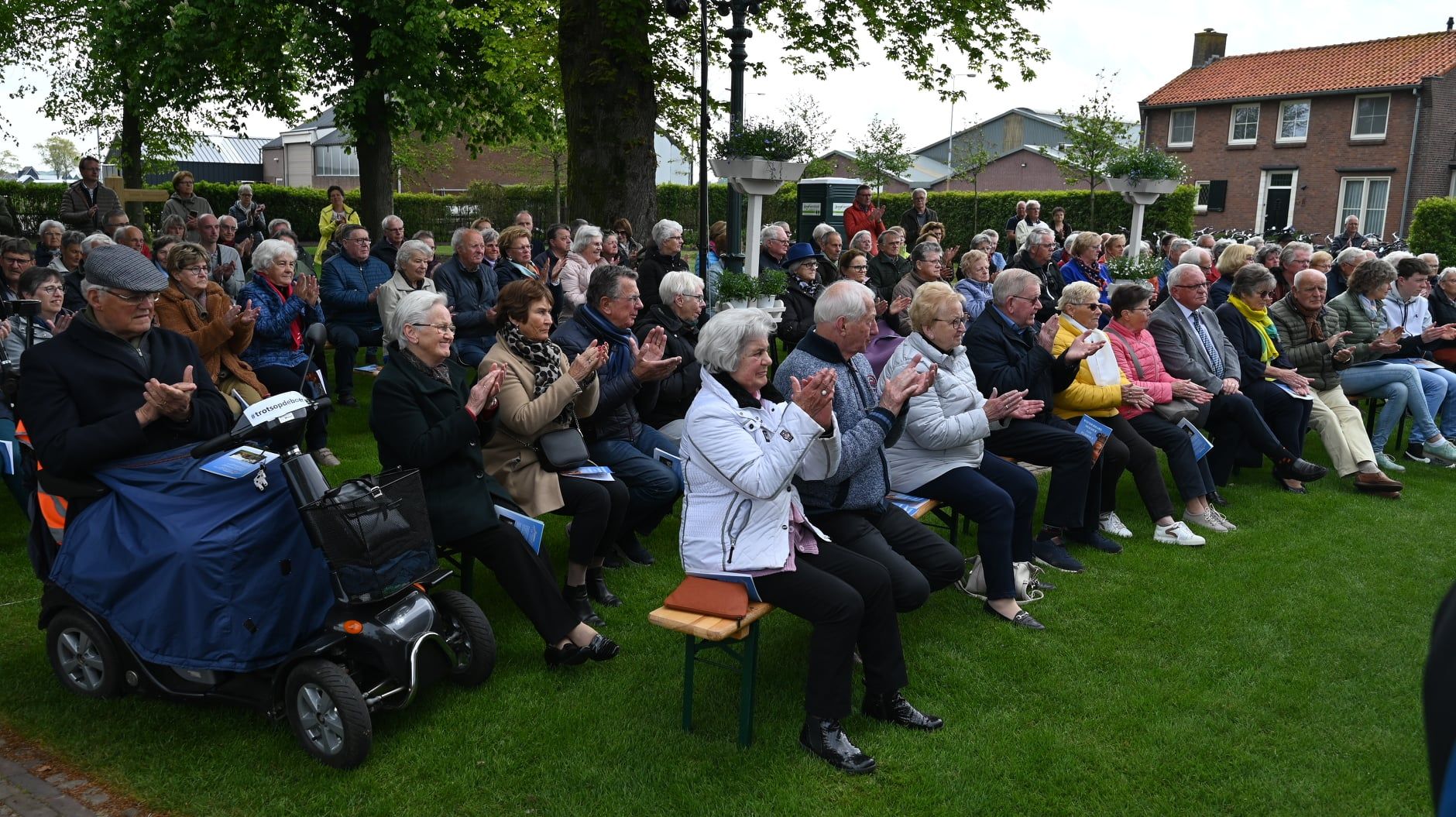 Publiek applaudisseert buiten; zittend op het gras. Mensen van verschillende leeftijden, gebouwen op de achtergrond.