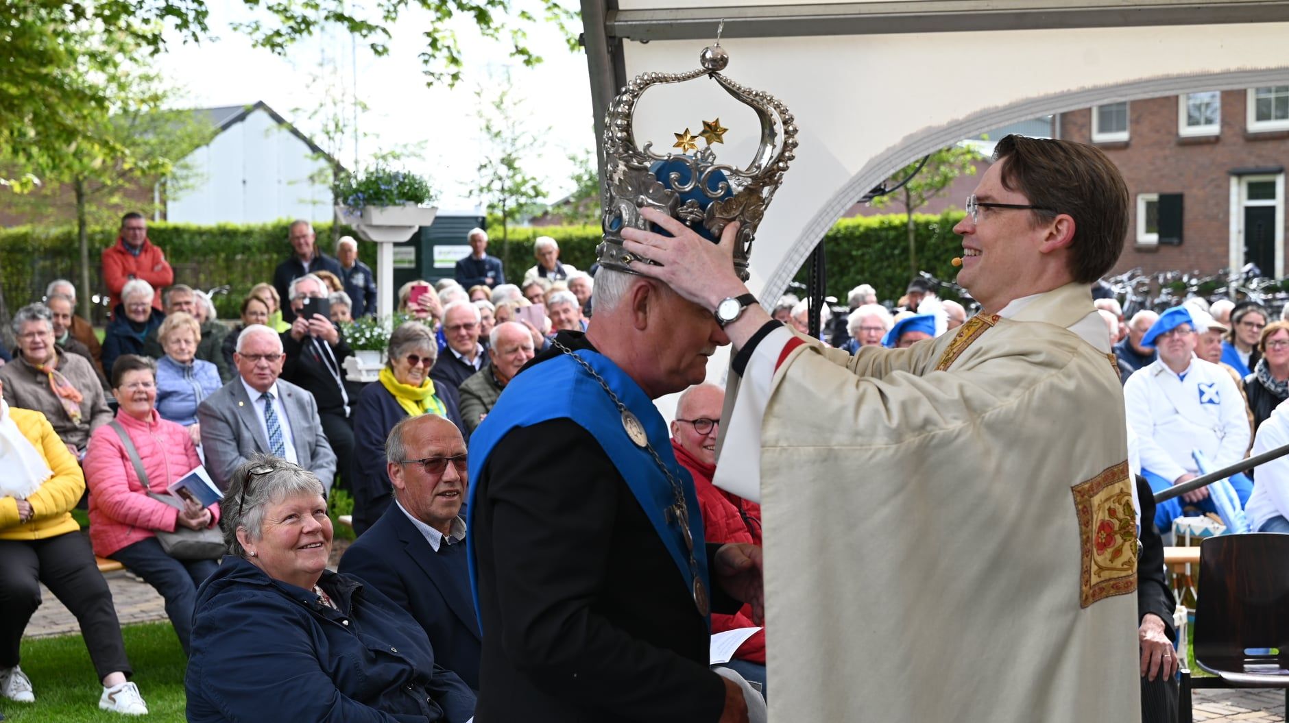 Een man wordt in de open lucht gekroond door een andere man in religieuze gewaden; een menigte kijkt toe.
