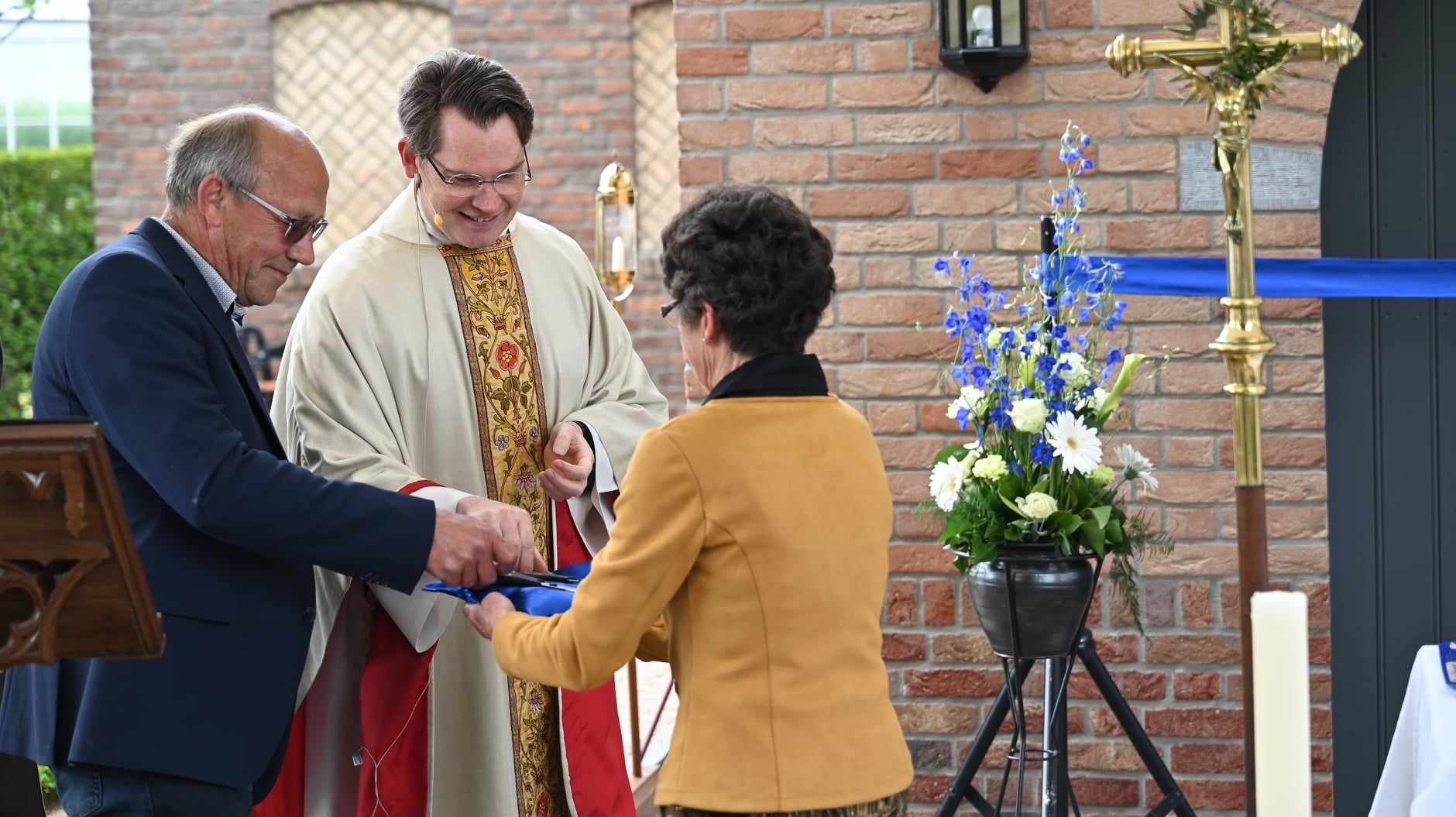 Mensen knippen een blauw lint door voor een bakstenen gebouw. ​​Een priester glimlacht terwijl hij het lint vasthoudt.