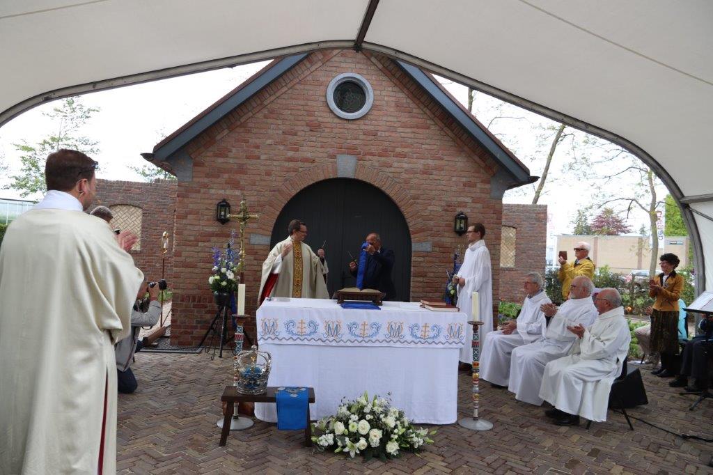 Religieuze ceremonie buiten een bakstenen gebouw. ​​Priesters in witte gewaden staan ​​rond een tafel onder een wit baldakijn.