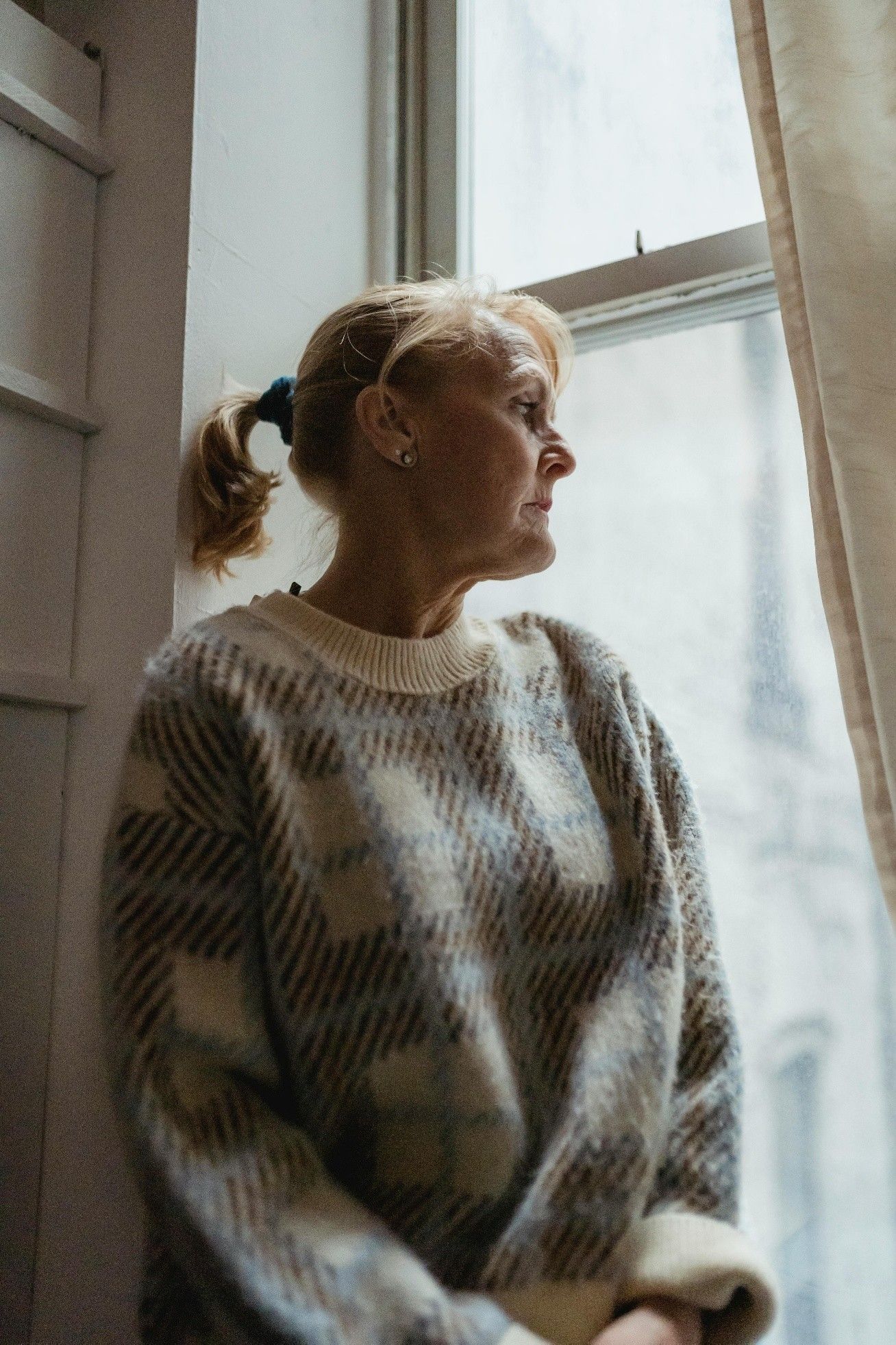Een vrouw met een blonde paardenstaart kijkt uit het raam. Ze draagt een trui met een patroon. Het licht stroomt naar binnen.