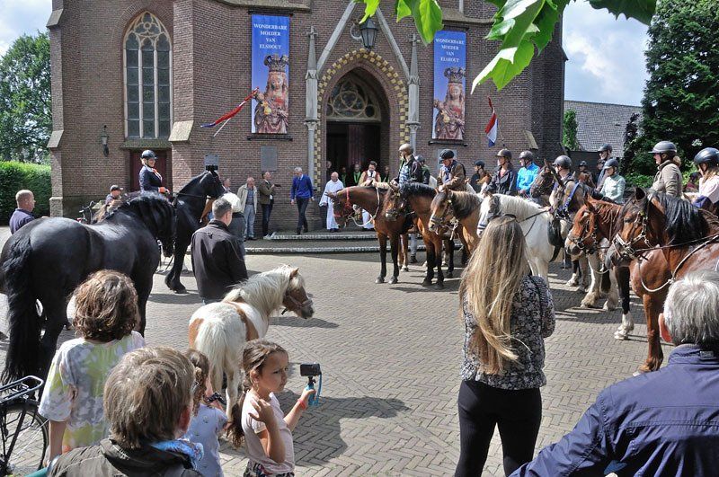 Mensen en paarden verzamelden zich voor een kerk. Ruiters te paard, toeschouwers keken toe. Blauwe vlaggen.