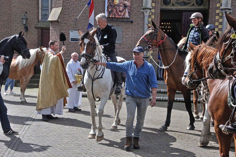 Paarden en ruiters voor een gebouw; een priester zegent een paard terwijl een man een ander paard leidt.