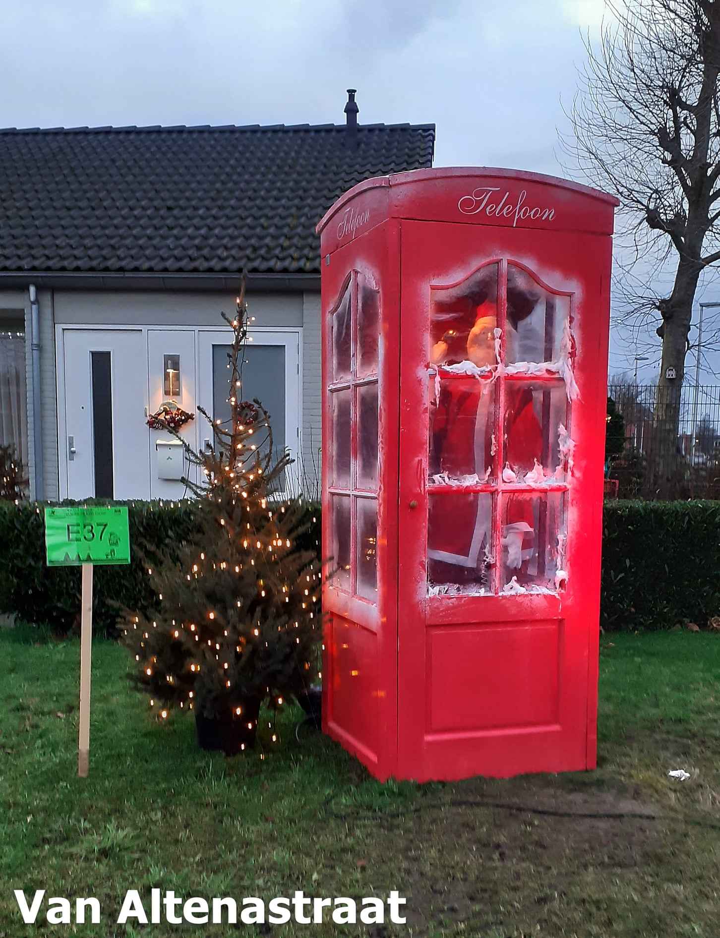 Een rode telefooncel met kerstverlichting en een kleine kerstboom in een tuin.