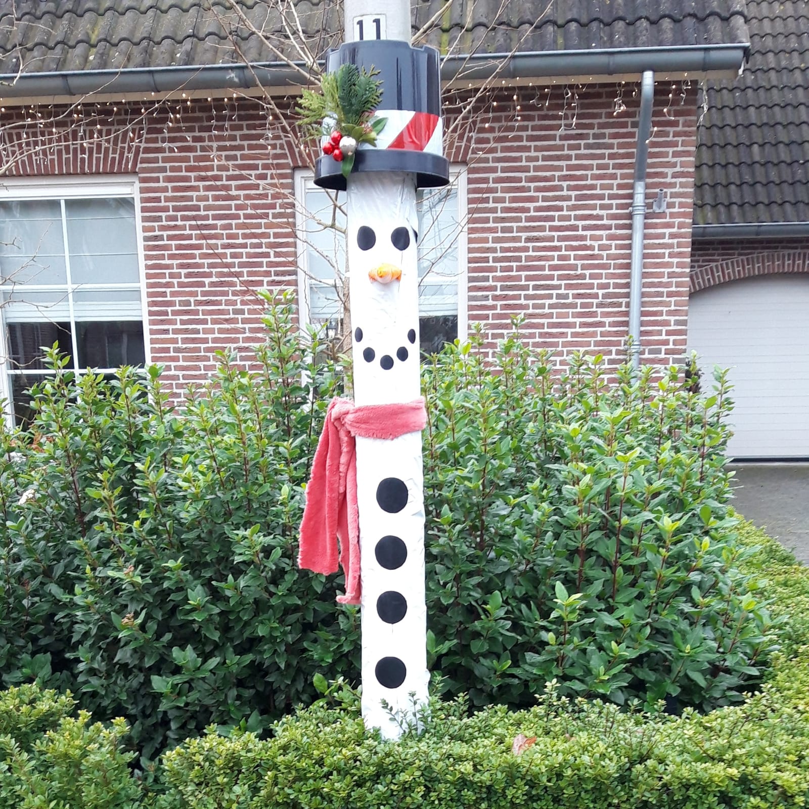 Een beschilderde paal, versierd als een sneeuwpop met een rode sjaal, een zwarte hoed en een bladerkrans, staat voor een bakstenen huis.