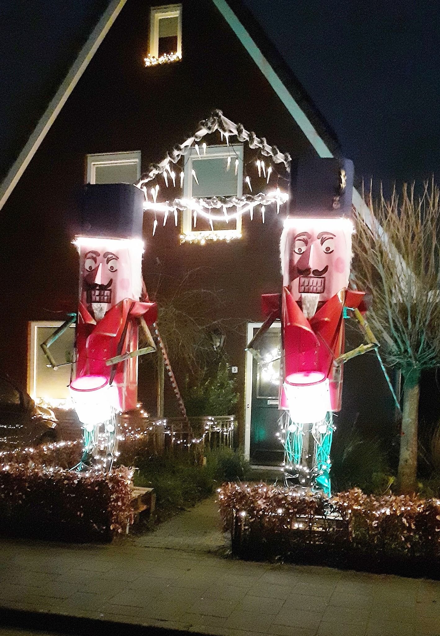 Twee verlichte, grote notenkrakerdecoraties flankeren de ingang van een huis, dat 's avonds is versierd met kerstverlichting.