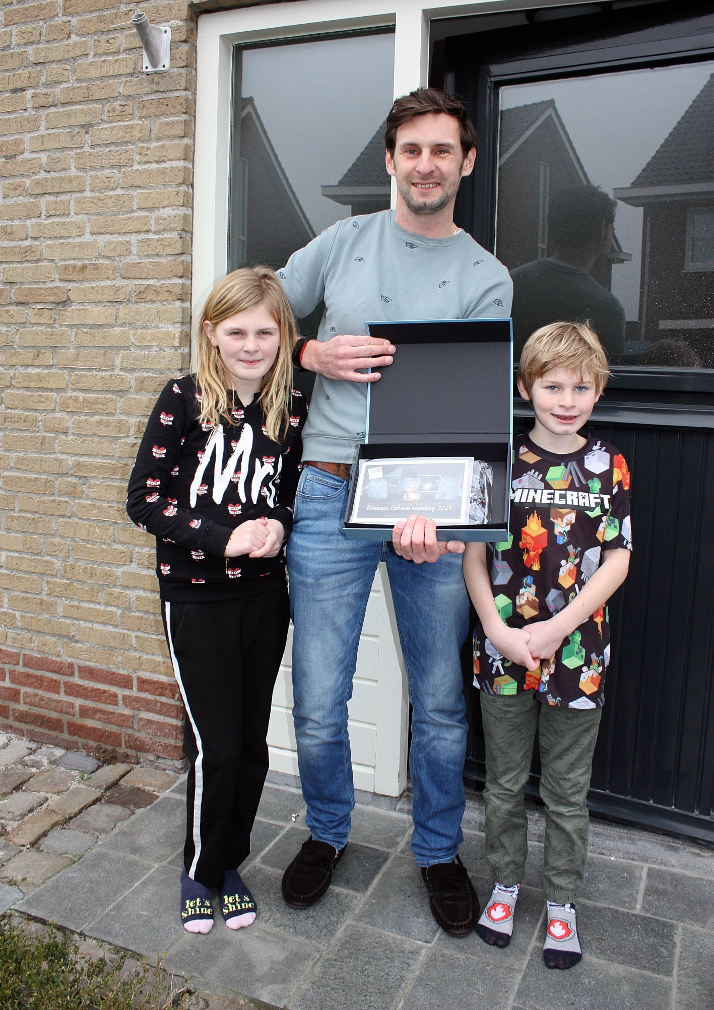 Een man en twee kinderen houden een zwarte doos vast voor een huis; de man lacht, de kinderen kijken naar de camera.