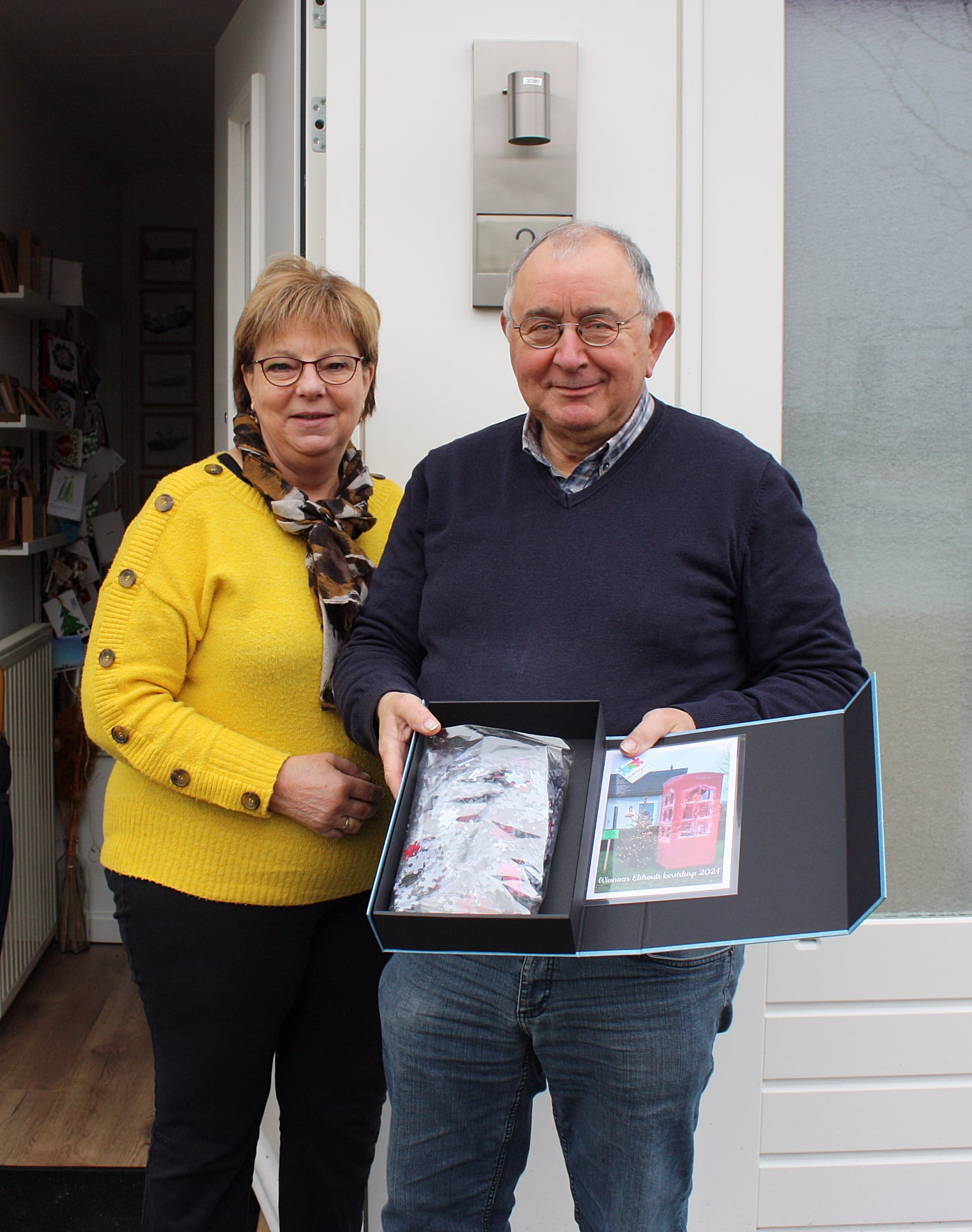 Een vrouw en een man staan glimlachend voor een deur, terwijl ze een open doos met kunstwerken vasthouden.