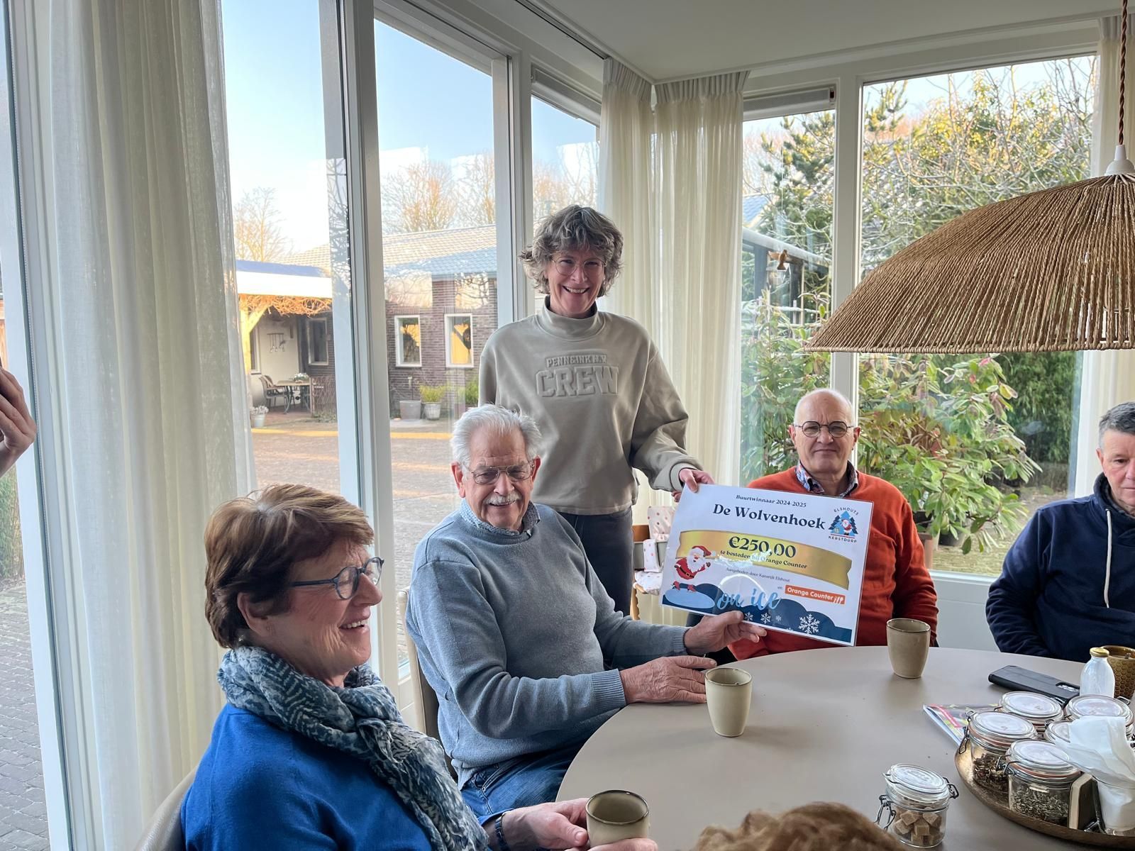Mensen zaten rond een tafel; iemand hield een certificaat vast. Het zonlicht stroomde naar binnen.