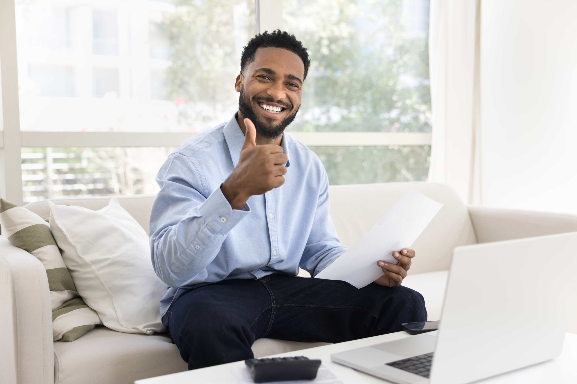 Person smiling and giving a thumbs-up while sitting on a couch with a laptop in a bright living room.