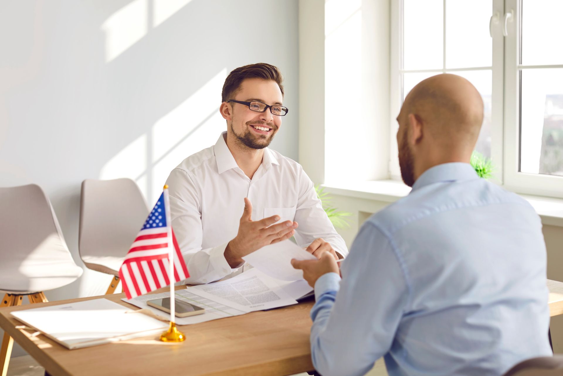 Two men talking at a desk in a bright office, with a small American flag between them.
